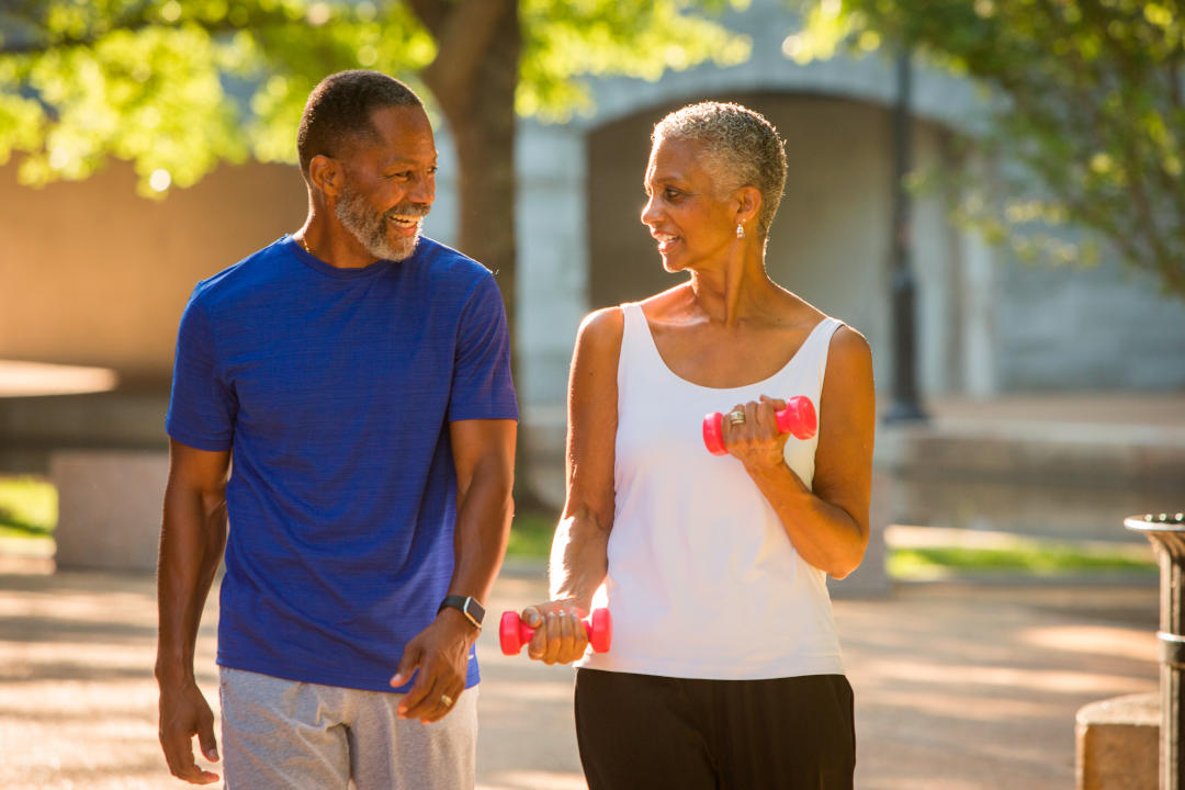 An older man with a blue shirt and a woman with a white tank top with dumbbells in her hands exercising outdoors 